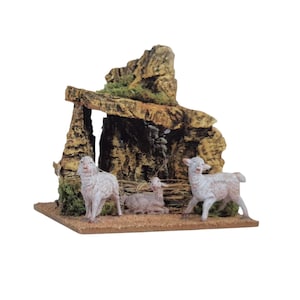 Può includere: Un presepe in miniatura con una struttura in pietra con un tetto muschioso e tre pecore bianche. Le pecore sono in varie pose, una sdraiata. La scena è ambientata su una base marrone.