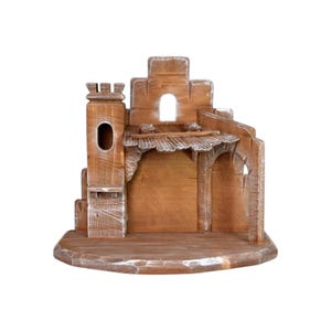 Può includere: Presepe in legno dal design rustico. La struttura presenta una torre, aperture ad arco e una zona coperta. Il legno ha una finitura naturale con accenti bianchi, creando un aspetto invecchiato. La base è rotonda.