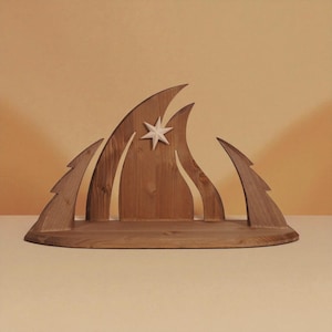 Può includere: Decorazione natalizia in legno con una stella e forme a fiamma stilizzate. Il pezzo in legno naturale include una stella bianca e due ritagli a forma di albero, il tutto su una base curva.