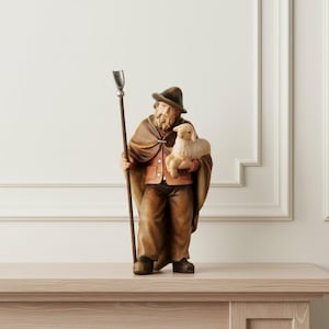 Op de afbeelding: Een handbeschilderd beeldje van een herder met baard, een bruine hoed, een beige cape en een bruine broek. Het figuur houdt een hoge houten staf met een zilverkleurige metalen punt in de ene hand en draagt een klein, crèmekleurig lammetje.