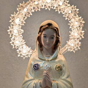 Peut inclure: Statue religieuse d'une femme en robe cr&egrave;me, les mains jointes en pri&egrave;re. Un halo de lumi&egrave;res en forme d'&eacute;toiles entoure sa t&ecirc;te. La robe est orn&eacute;e d'ornements en forme de roses pourpres, blanches et dor&eacute;es.