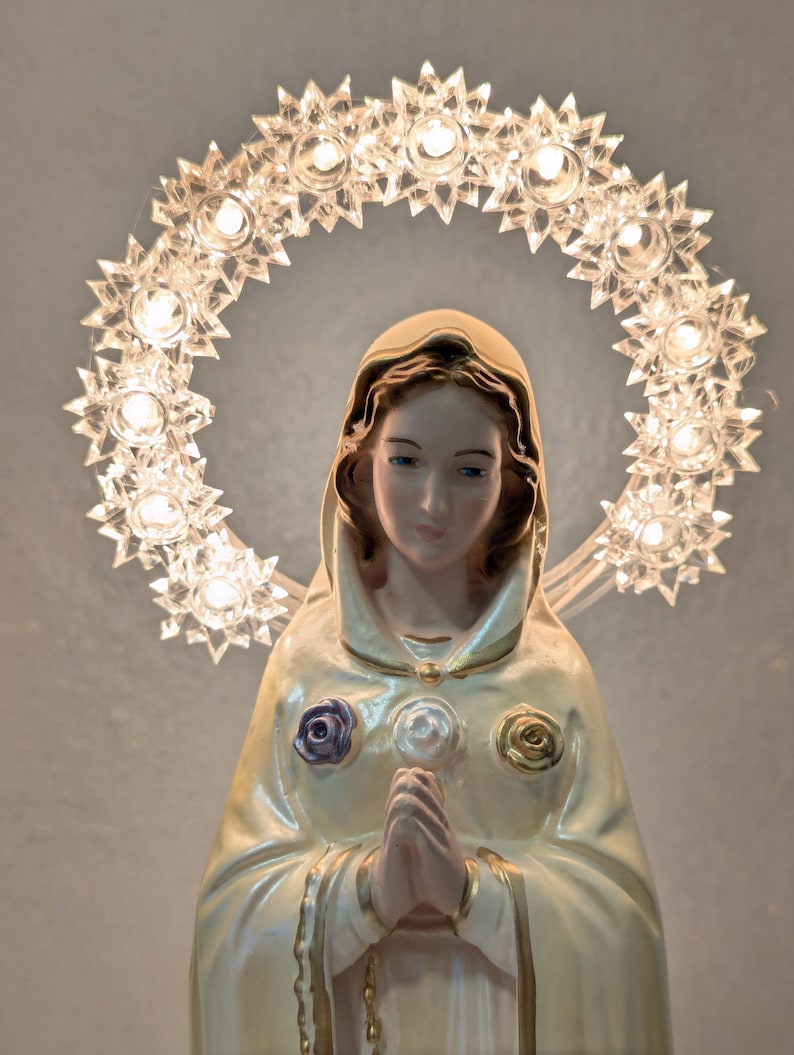 Peut inclure: Statue religieuse d'une femme en robe cr&egrave;me, les mains jointes en pri&egrave;re. Un halo de lumi&egrave;res en forme d'&eacute;toiles entoure sa t&ecirc;te. La robe est orn&eacute;e d'ornements en forme de roses pourpres, blanches et dor&eacute;es.