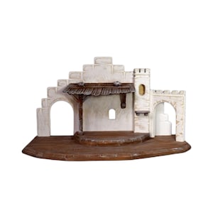 Può includere: Un presepe dettagliato, bianco sporco, con una base in legno marrone. La scena presenta una struttura centrale con tetto di paglia, archi e una piccola torre. Il design generale ricorda un ambiente rustico mediorientale.