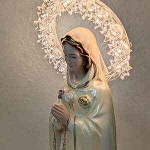 Peut inclure: Statuette religieuse d'une femme en robe cr&egrave;me, tenant une fleur. Un halo de cristaux clairs et facett&eacute;s entoure sa t&ecirc;te. La figurine a une expression sereine et est orn&eacute;e d'accents dor&eacute;s.
