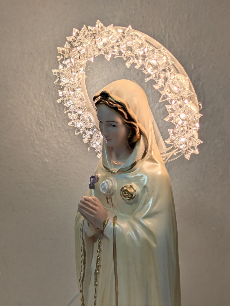 Peut inclure: Statuette religieuse d'une femme en robe cr&egrave;me, tenant une fleur. Un halo de cristaux clairs et facett&eacute;s entoure sa t&ecirc;te. La figurine a une expression sereine et est orn&eacute;e d'accents dor&eacute;s.