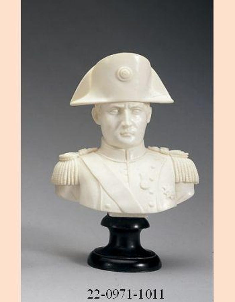 May include: A white ivory bust sculpture of a man wearing a military uniform and a bicorne hat. The bust is on a black wooden base.