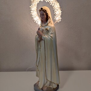Peut inclure: Statue cr&egrave;me d'une femme en robe fluide, tenant une fleur et un chapelet. Un halo de lumi&egrave;res en forme d'&eacute;toiles entoure sa t&ecirc;te. La statue est sur une base rectangulaire grise.