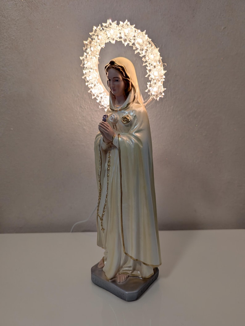 Peut inclure: Statue cr&egrave;me d'une femme en robe fluide, tenant une fleur et un chapelet. Un halo de lumi&egrave;res en forme d'&eacute;toiles entoure sa t&ecirc;te. La statue est sur une base rectangulaire grise.