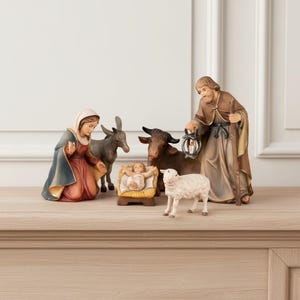 Op de afbeelding: Een kerststal-figurenset met een knielende figuur in een blauw en rood gewaad, een staande figuur in een bruin gewaad met een lantaarn en staf, een baby in een kribbe, een ezel, een bruine os en een wit schaap, allemaal in een realistische stijl.