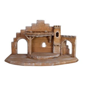Può includere: Un presepe in legno dal design rustico. La struttura presenta archi, una piccola torre e una zona coperta. Il colore è marrone chiaro con accenti bianchi, creando un aspetto invecchiato. La scena è progettata per contenere statuine.
