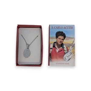 May include: A stainless steel necklace with an oval pendant, displayed in a red box. The box also features the text "STAINLESS STEEL". Beside the box is a card with an image of a young man and the text "S. CARLO ACUTIS" and "STAINLESS STEEL".
