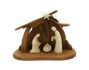 Può includere: Un presepe in legno con una stalla in legno marrone e tre figure bianche che rappresentano Maria, Giuseppe e il bambino Gesù. La stalla ha una stella in cima.