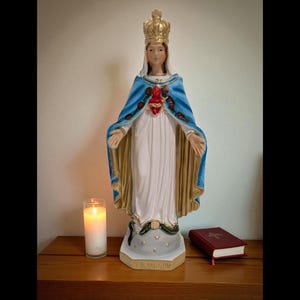 May include: A religious statue of a woman wearing a crown and blue cape with gold trim. The statue has a red heart on the chest and is standing on a base with stars. A lit candle and a red book are on a wooden surface.