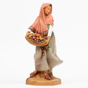 May include: A hand-painted figurine of a person wearing a peach-coloured headscarf and a long, flowing grey skirt. The figure holds a woven basket filled with various fruits, including apples and pears, and wears brown shoes on a base.
