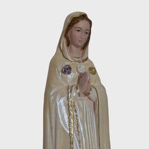 Peut inclure: Statue cr&egrave;me d'une figure v&ecirc;tue d'une robe, les mains jointes en pri&egrave;re. La figure porte un manteau &agrave; capuche orn&eacute; d'une bordure dor&eacute;e et de trois ornements en forme de rose. Un chapelet dor&eacute; pend devant.