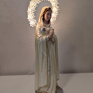 Peut inclure: Statue religieuse d'une femme v&ecirc;tue d'une robe cr&egrave;me, avec un halo de lumi&egrave;res. La statue a une expression sereine et joint les mains en pri&egrave;re. Une cha&icirc;ne de perles pend de son cou. La base est un carr&eacute; argent&eacute;.