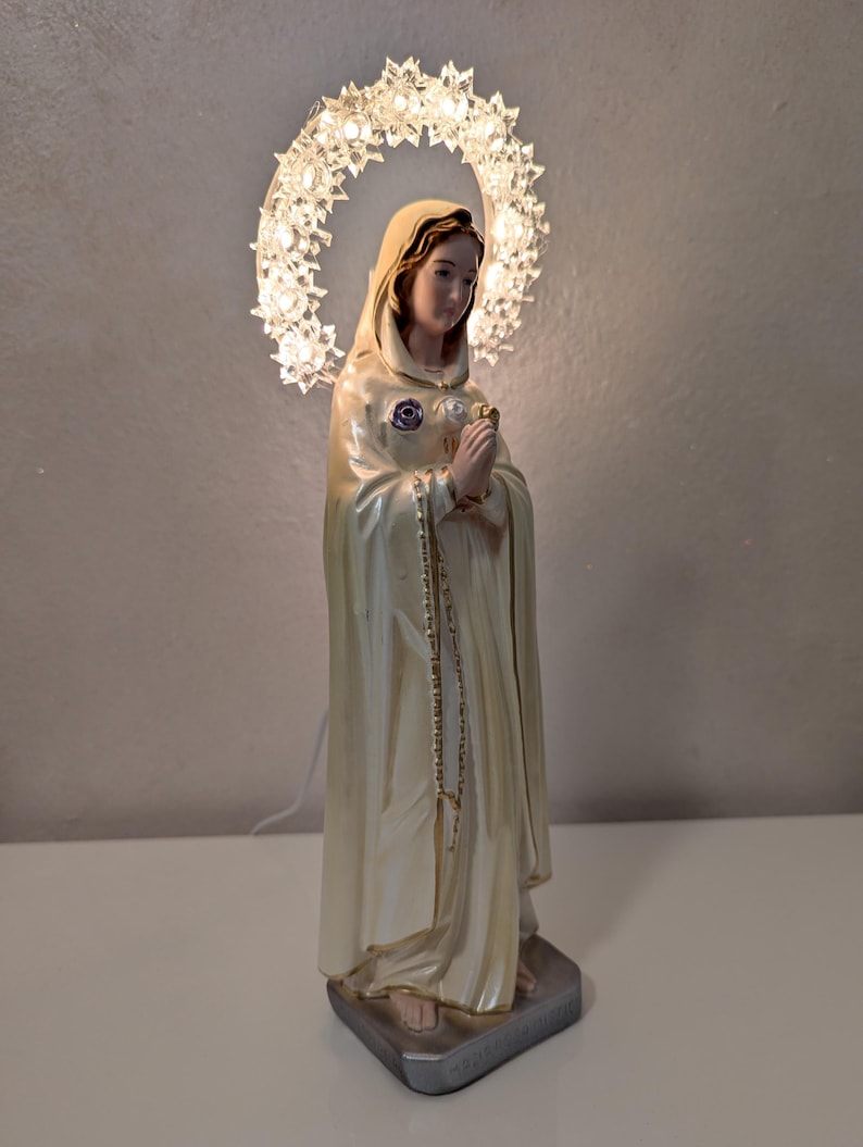 Peut inclure: Statue religieuse d'une femme v&ecirc;tue d'une robe cr&egrave;me, avec un halo de lumi&egrave;res. La statue a une expression sereine et joint les mains en pri&egrave;re. Une cha&icirc;ne de perles pend de son cou. La base est un carr&eacute; argent&eacute;.