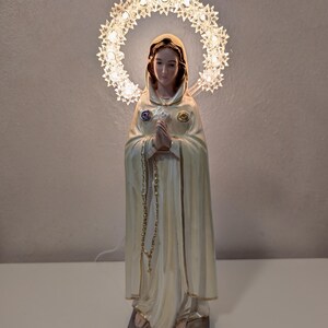 Peut inclure: Statue religieuse d'une femme en robe cr&egrave;me, les mains jointes en pri&egrave;re. Un halo de petites lumi&egrave;res illumin&eacute;es entoure sa t&ecirc;te. La statue a un chapelet dor&eacute; et est mont&eacute;e sur une base rectangulaire.