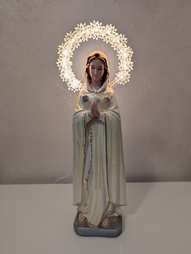 Peut inclure: Statue religieuse d'une femme en robe cr&egrave;me, les mains jointes en pri&egrave;re. Un halo de petites lumi&egrave;res illumin&eacute;es entoure sa t&ecirc;te. La statue a un chapelet dor&eacute; et est mont&eacute;e sur une base rectangulaire.