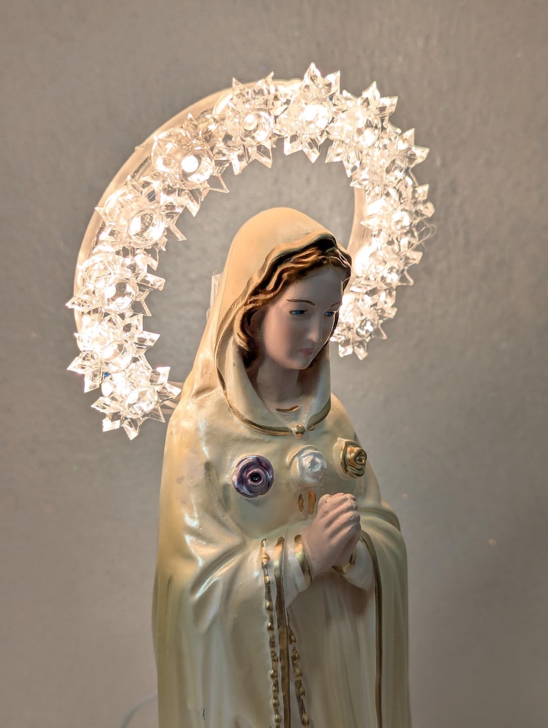 Peut inclure: Figurine religieuse d'une femme en robe cr&egrave;me, avec un halo de lumi&egrave;res en forme d'&eacute;toiles. La figurine a une expression sereine et est orn&eacute;e d'ornements en forme de roses. Un chapelet est suspendu au cou de la figurine.