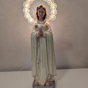 Peut inclure: Statue cr&egrave;me d'une femme en cape &agrave; capuche, les mains jointes en pri&egrave;re. Un halo de petites lumi&egrave;res en forme d'&eacute;toiles illumine sa t&ecirc;te. La statue repose sur un socle rectangulaire.