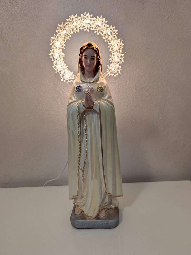 Peut inclure: Statue cr&egrave;me d'une femme en cape &agrave; capuche, les mains jointes en pri&egrave;re. Un halo de petites lumi&egrave;res en forme d'&eacute;toiles illumine sa t&ecirc;te. La statue repose sur un socle rectangulaire.