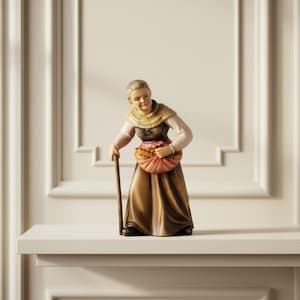 Op de afbeelding: Een handbeschilderd houten beeldje van een ouder persoon met grijs haar, gekleed in een beige sjaal, een donker lijfje en een lange bruine rok. Het figuur houdt een houten wandelstok en een stoffen bundel met brood in de handen.