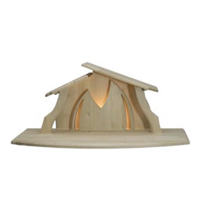Può includere: Un presepe in legno con luce interna. La struttura ha un tetto e aperture ad arco. La base è una superficie piana in legno. Il legno è di colore chiaro naturale.