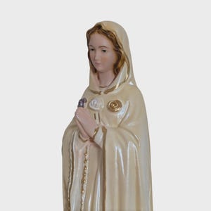 Peut inclure: Statue cr&egrave;me d'une femme en cape &agrave; capuche, avec garnitures et accents dor&eacute;s. La statue a une expression sereine et les mains jointes, avec des d&eacute;tails de roses sur la cape. Un petit chapelet est suspendu &agrave; la cape.