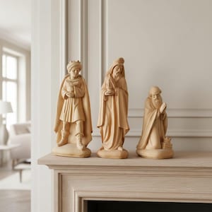 Op de afbeelding: Drie lichtgekleurde houten beeldjes van figuren in gewaden en hoofddeksels. De linkerfiguur draagt een tulband, de middelste figuur houdt een voorwerp vast en de rechterfiguur knielt met gevouwen handen, allen op ovale voetstukken.