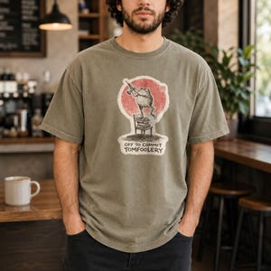 May include: An olive green short-sleeve t-shirt featuring a graphic of a frog standing on a stack of books on a chair, holding a drill. Behind the frog is a red circle. The text below reads: OFF TO COMMIT TOMFOOLERY. The shirt has a crew neckline.
