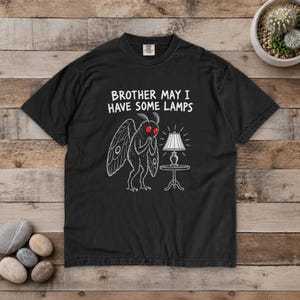 May include: A black t-shirt featuring a white line art illustration of a moth with bright red eyes standing next to a lamp on a table. The text above the moth reads, "BROTHER MAY I HAVE SOME LAMPS" in white capital letters.