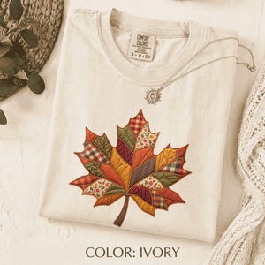May include: An ivory short-sleeved t-shirt featuring a large, central maple leaf graphic composed of various quilted fabric patterns in shades of red, orange, yellow, green, and brown. The shirt has a crew neckline and a Comfort Colors brand label.