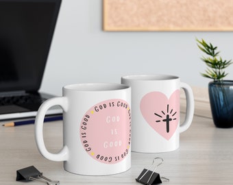 Op de afbeelding: Twee witte keramische mokken met roze accenten. Eén mok heeft de tekst "God is Good" in een cirkel met gele stippen. De andere mok heeft een roze hart met een kruis erin.