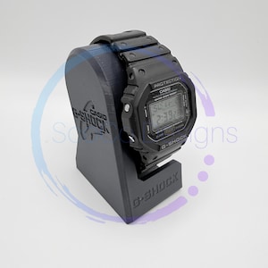 May include: A black G-Shock watch on a black stand. The watch face displays the time. The stand features the Casio G-Shock logo. The watch is a digital watch with a black strap. The stand is angled.