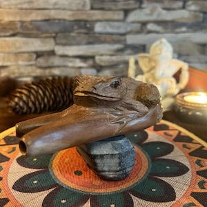 Può includere: Una pipa di legno intagliata a mano a forma di rana, appoggiata su una piccola pietra. La pipa è marrone con intagli dettagliati di una rana. Lo sfondo include un tappetino decorativo, una pigna e una candela accesa.