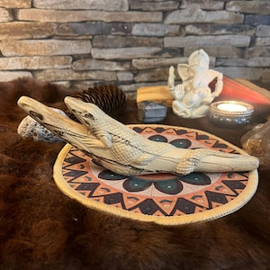 Può includere: Una statuetta di alligatore in legno intagliato poggia su un tappetino circolare decorativo con un motivo geometrico. L'alligatore è marrone chiaro con segni marrone scuro. Il tappetino è su una superficie marrone, con un muro di pietra sullo sfondo.