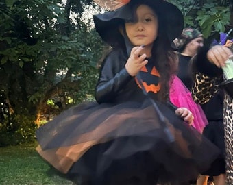 May include: A young girl dressed as a witch for Halloween. She is wearing a black dress with a tulle skirt, a black witch hat with an orange and black ribbon, and black boots. She is standing in a grassy area with a red plastic slide in the background.