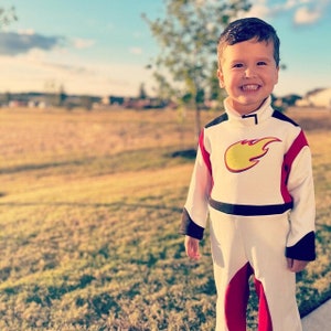 May include: A young boy dressed in a white, red, and black astronaut costume with a yellow flame design on the chest. He is smiling and standing in a grassy field.