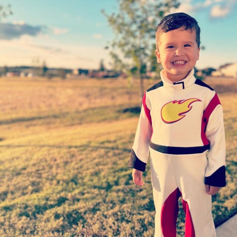 May include: A young boy dressed in a white, red, and black astronaut costume with a yellow flame design on the chest. He is smiling and standing in a grassy field.