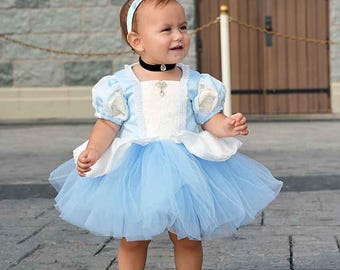 Könnte beinhalten: Ein Baby in einem hellblauen Kleid im Cinderella-Stil mit passendem Stirnband und weißen Schuhen. Das Kleid hat einen Tüllrock und Puffärmel. Das Baby trägt eine schwarze Halskette mit Anhänger.