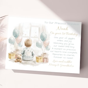 May include: A watercolor illustration of a young boy looking out a window with balloons and presents. The text reads "To Our Precious Grandson Noah On your 1st Birthday One year of giggles, smiles, and joy. Watching you grow, our sweet little boy. A moment so precious, our hearts now complete. Memories to cherish forever so sweet. Love and cuddles Nanny & Granddad xx"