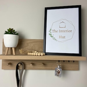 May include: A wooden shelf with hooks and a small shelf on top. The shelf is mounted on a white wall. There is a potted plant, a string of wooden beads, and a framed print with the text "The Interior Hut" on the shelf. A key chain with a photo of a dog is hanging from one of the hooks.