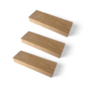 Op de afbeelding: Drie rechthoekige houten blokken met een lichtbruine afwerking zijn diagonaal geplaatst tegen een witte achtergrond. De blokken hebben een glad, schoon uiterlijk en tonen de natuurlijke houtnerf. De blokken zijn waarschijnlijk voor interieurdecoratie of knutselen.