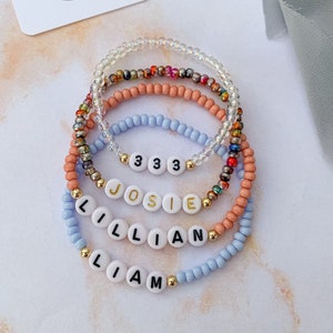 May include: Four beaded bracelets with white beads and gold lettering. The bracelets spell out the names "Liam", "Lillian", "Josie", and the number "333".