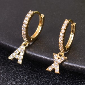 May include: A pair of gold hoop earrings with a dangling letter charm. The earrings are set with clear cubic zirconia stones. The letter charms are 'A' and 'X'.