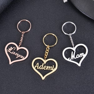Könnte beinhalten: Drei herzförmige Schlüsselanhänger mit verschiedenen Namen darauf. Die Schlüsselanhänger sind in Gold, Silber und Roségold. Die Namen auf den Schlüsselanhängern sind "Love You", "Mom" und "Ademi".