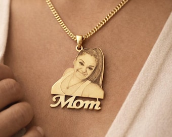 Può includere: Collana color oro con un ciondolo con un ritratto e la parola "Mom". Il ciondolo è rettangolare con un ritaglio del volto di una donna e la parola "Mom" in corsivo sotto. La catena è a maglia a catena.