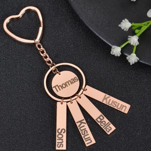 May include: Rose gold keychain with a heart-shaped ring and five dangling tags. The tags are engraved with the names "Thomas", "Kusun", "Bella", "Kusun", and "Sons".
