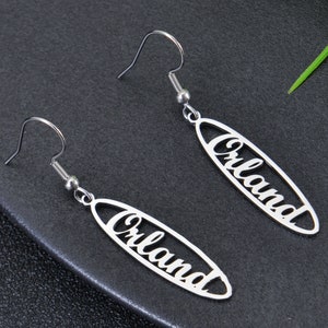 May include: A pair of silver dangle earrings with the word "Orlando" cut out in a script font. The earrings are oval shaped and have a hook fastening.